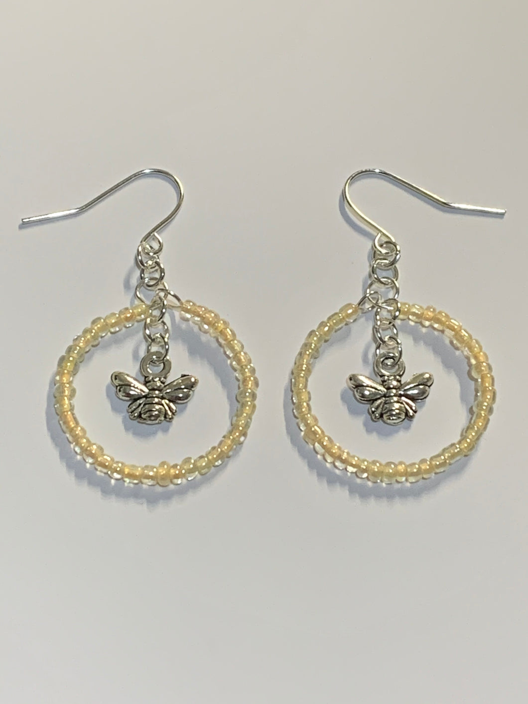 Silver coloured findings with a circle of yellow mini beads, framing a little bee charm in the centre. Earrings lay flat against a white background