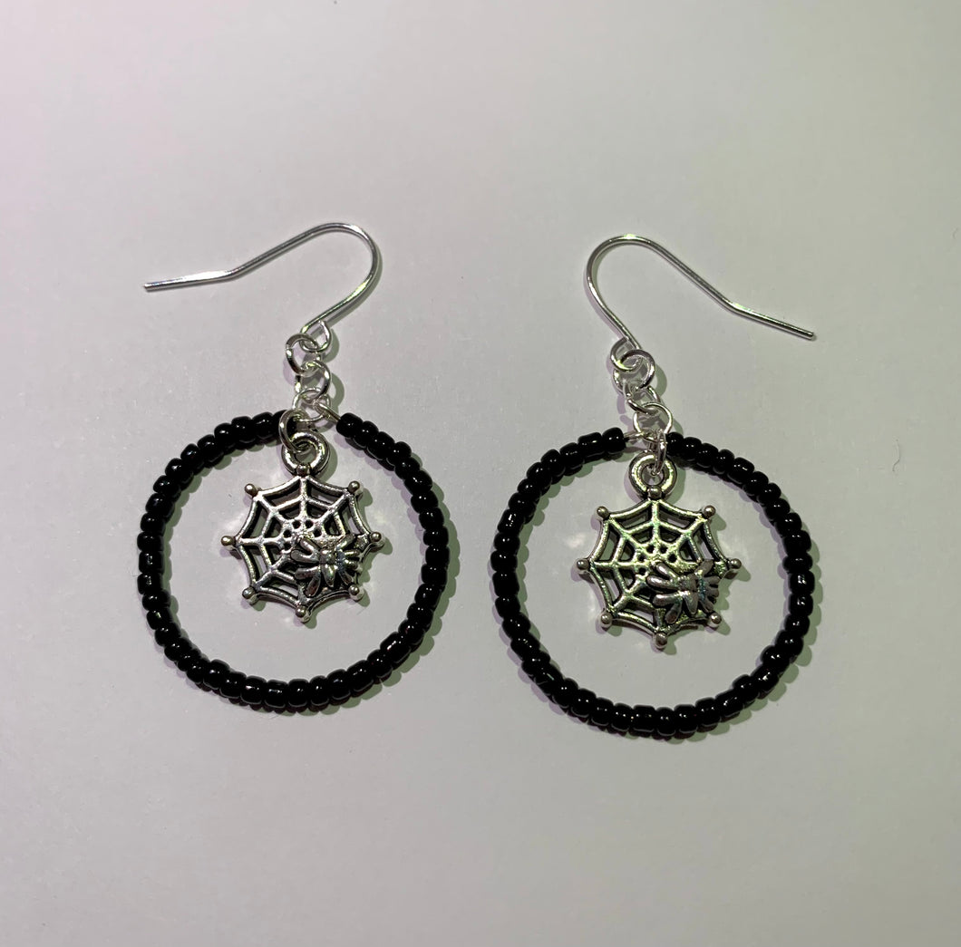 earrings laying flat againast white background. silver coloured hooks and charms. spiderweb charms hang in the middle of a hoop/circle of small black beads