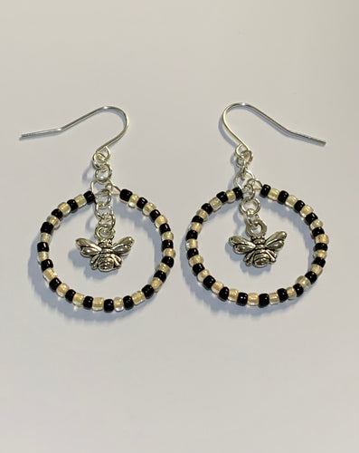 Silver coloured findings with a circle of alternate black and yellow mini beads, framing a little bee charm in the centre. Earrings lay flat against a white background