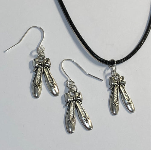 Image shows a pair of earrings laid flat against a white background, with part of a necklace in shot, showing the charm. Necklace is a black cord. Findings and charms are silver coloured. The charm is a pair of ballet slippers with a bow at the top, holding them both together.