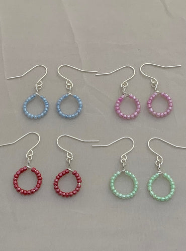 Image shows four pairs of earrings, each lay flat against a light grey background. Each have silver coloured findings and the beaded part is shaped into a small hoop/circle. Each pair has one colour of beads; blue, pink, green and cherry red